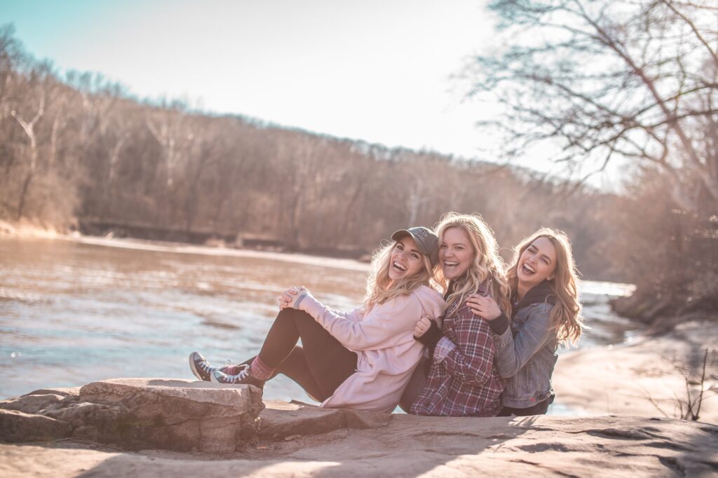 Three women sit close together outside by a river, smiling and laughing. They feel great after seeing Dr. Meena Malhotra, their provider at Heal n Cure wellness center in Glenview.
