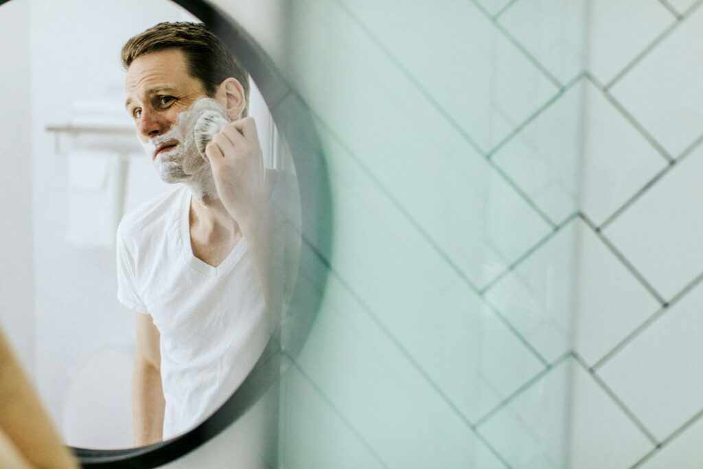 A man unhappily applies shaving cream to his face, contemplating laser hair removal for men in Glenview.